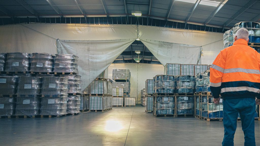 Image show a warehouse with pallets in it. There is a a dividing curtain and a man in a high visibility jacket in the foreground