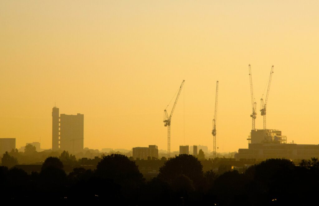 Image shows a silhouetted city skyline with cranes on a construction project, against an orange sunset.