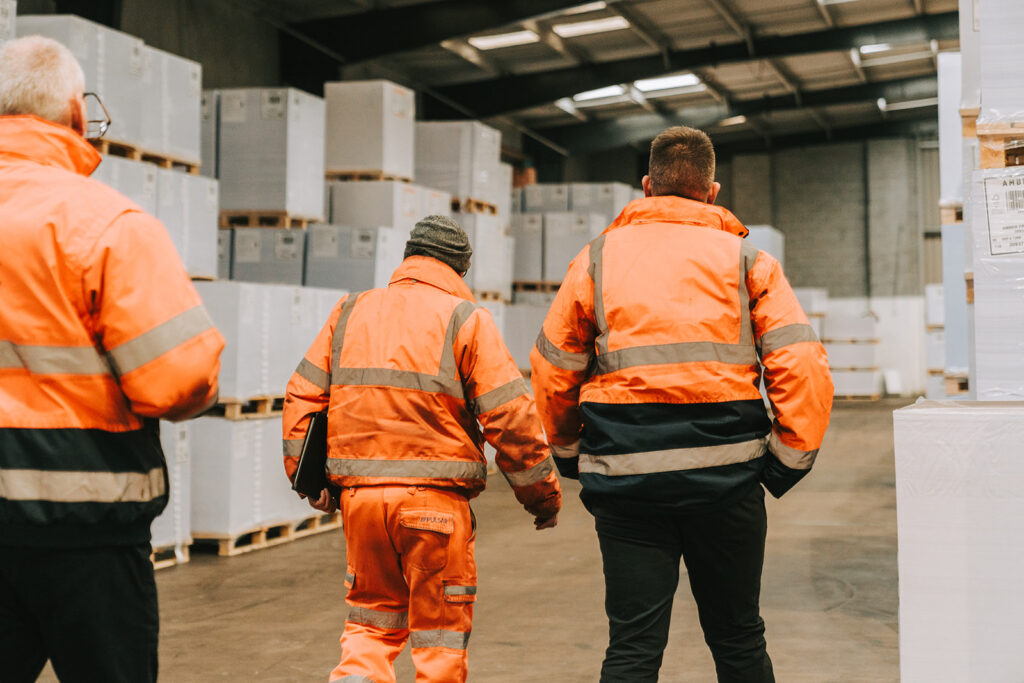 Shot shows three people in visibility clothing walking through a warehouse