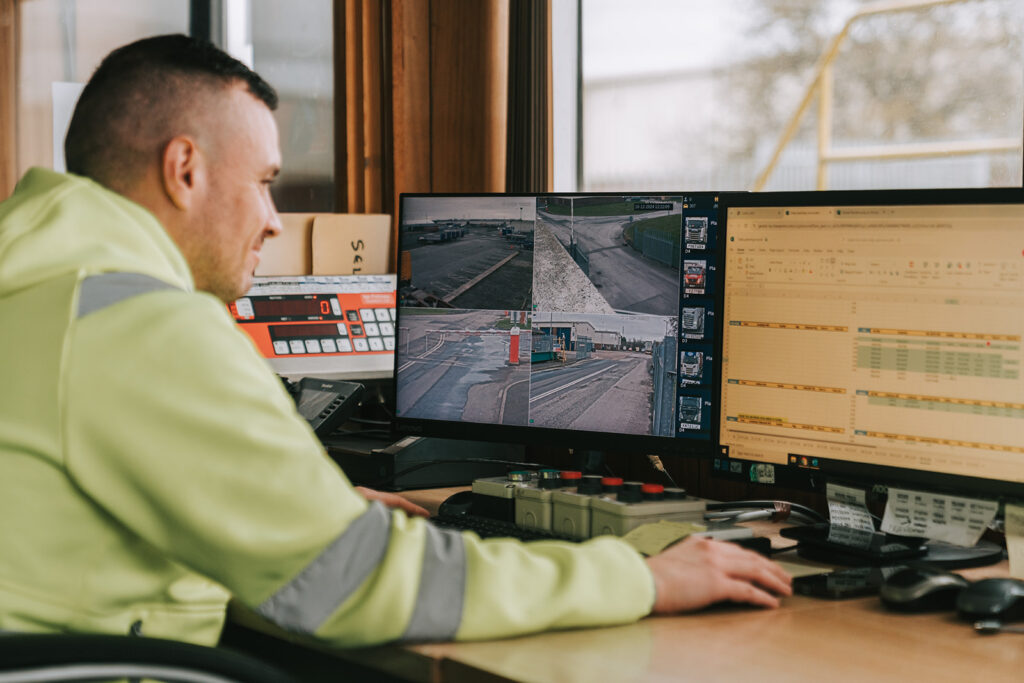 Image shows a man wearing a yellow high visibility jumper looking at a screen of CCTV images in a secure warehouse.