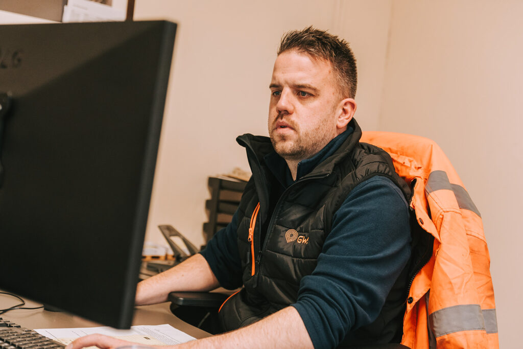 Image shows a man sitting at a desk looking at a computer screen in a warehouse.